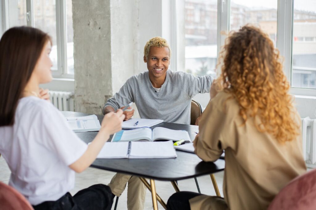 Positive African American male student and unrecognizable classmates looking at each other while sitting at table with textbooks and copybooks during lesson in classroom