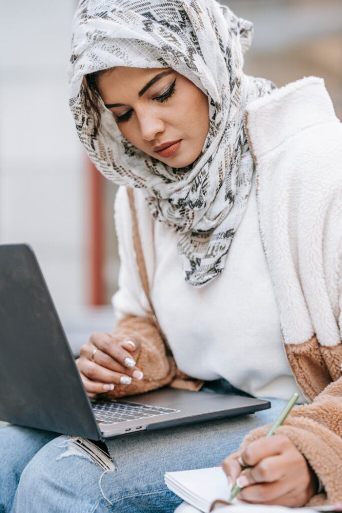 Young ethnic female freelancer in warm outfit and hijab using laptop while making notes in notebook with pen in daytime