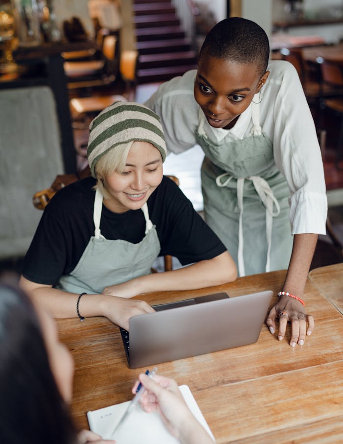 From above of optimistic diverse female workers of cafeteria discussing business plan while using laptop and taking notes in piece of paper at table