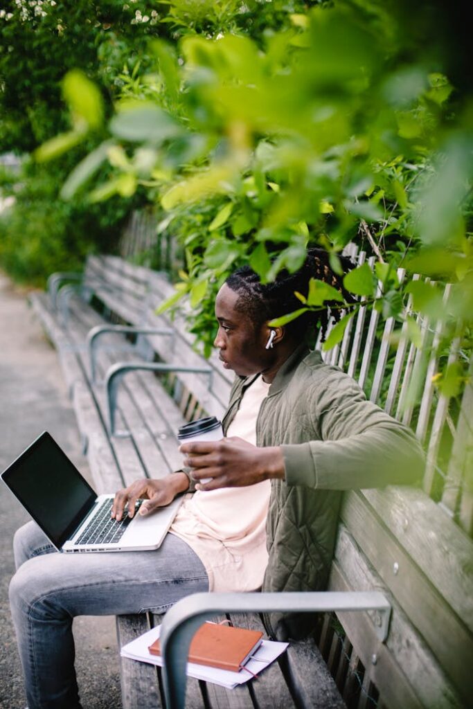 Side view of young African American male wearing wireless earbuds with coffee to go and laptop spacing out while sitting with notebooks on park bench in summer