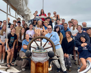 A large group of people pose together on a sailboat deck behind the ship’s wheel on a cloudy day.