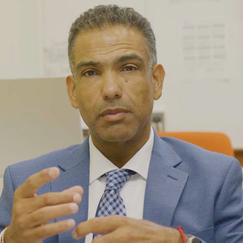 A man in a blue suit and checkered tie gestures while speaking in an indoor setting.