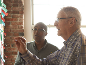 Two people collaborate by adding colorful sticky notes to a wall in a bright room with brick accents and large windows.