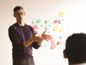 A person stands in front of a wall covered with colorful sticky notes, gesturing while speaking to someone seated in front of them.
