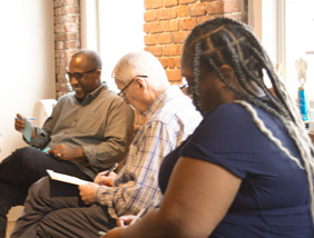 Three people sit by a window with brick walls, writing notes and smiling during a group activity.