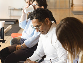 A group of people sit together in a bright room, writing notes and sharing ideas during a meeting.